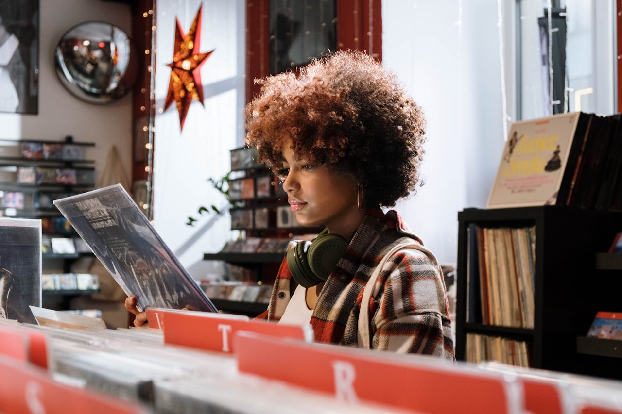 contact-header A woman with afro hair examines vinyl records in a cozy music shop, wearing headphones.